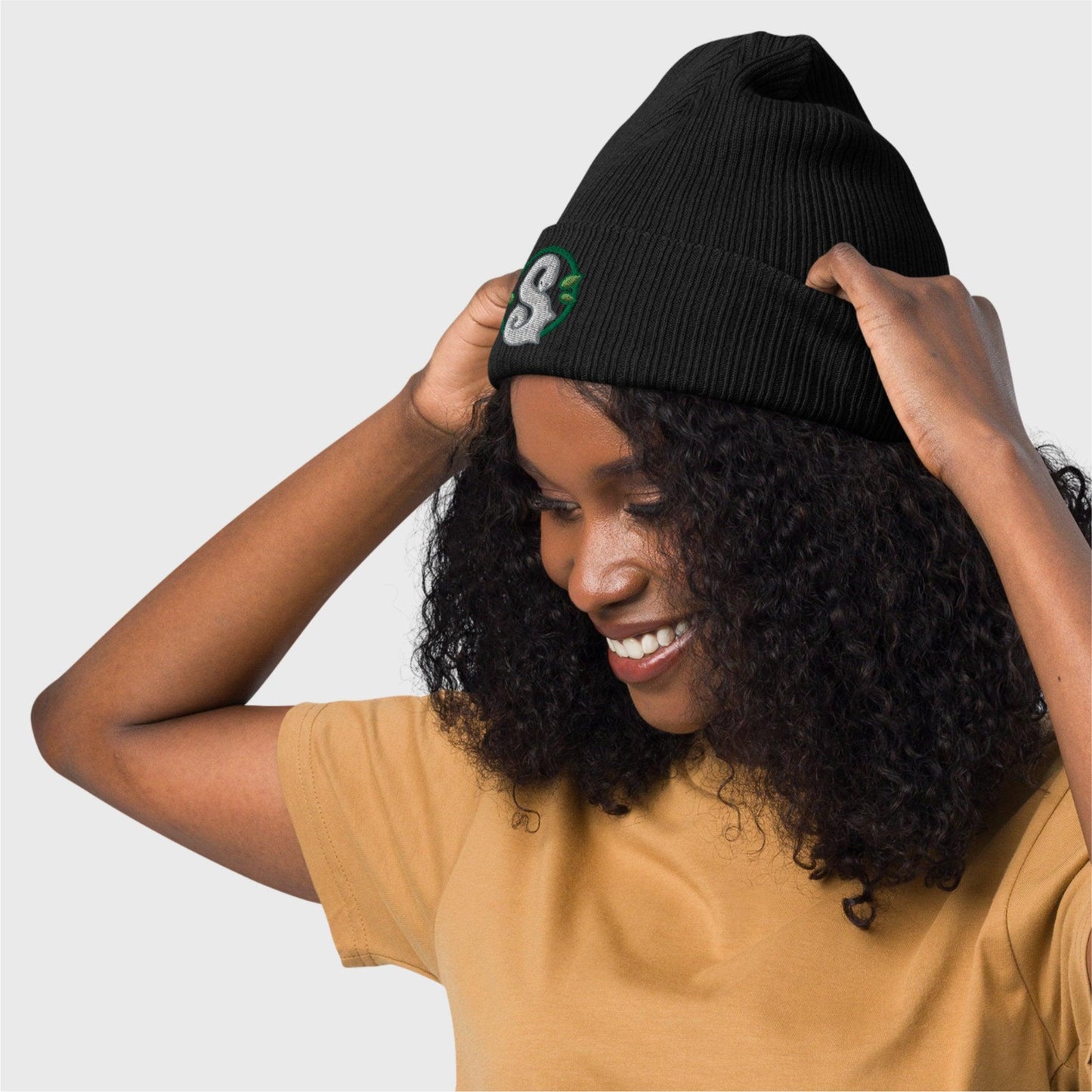 A woman smiles looking down on her right and holds the beanie back folds showcasing the embroidered logo on the front.