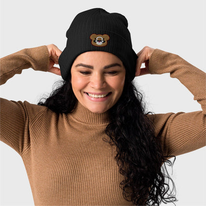 A woman holds smiles looking down and holds the beanie back folds showcasing the embroidered logo on the front.